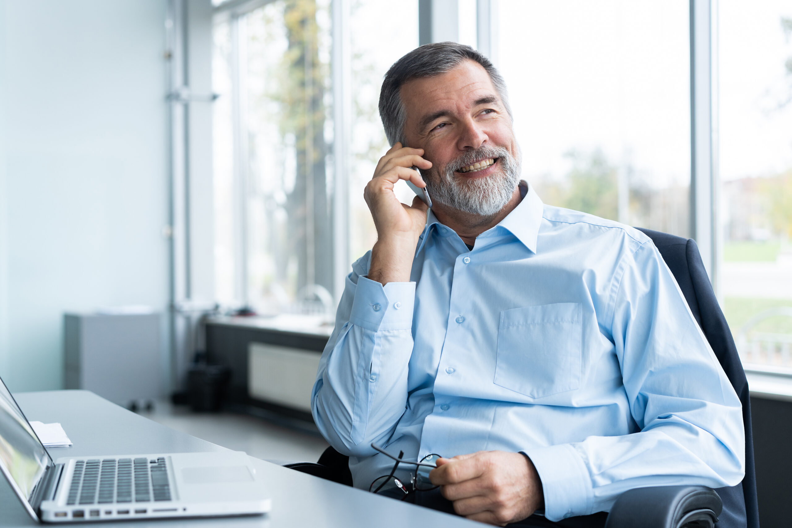 Executive senior businessman using his mobile phone and talking wih somebody while working laptop in the office.